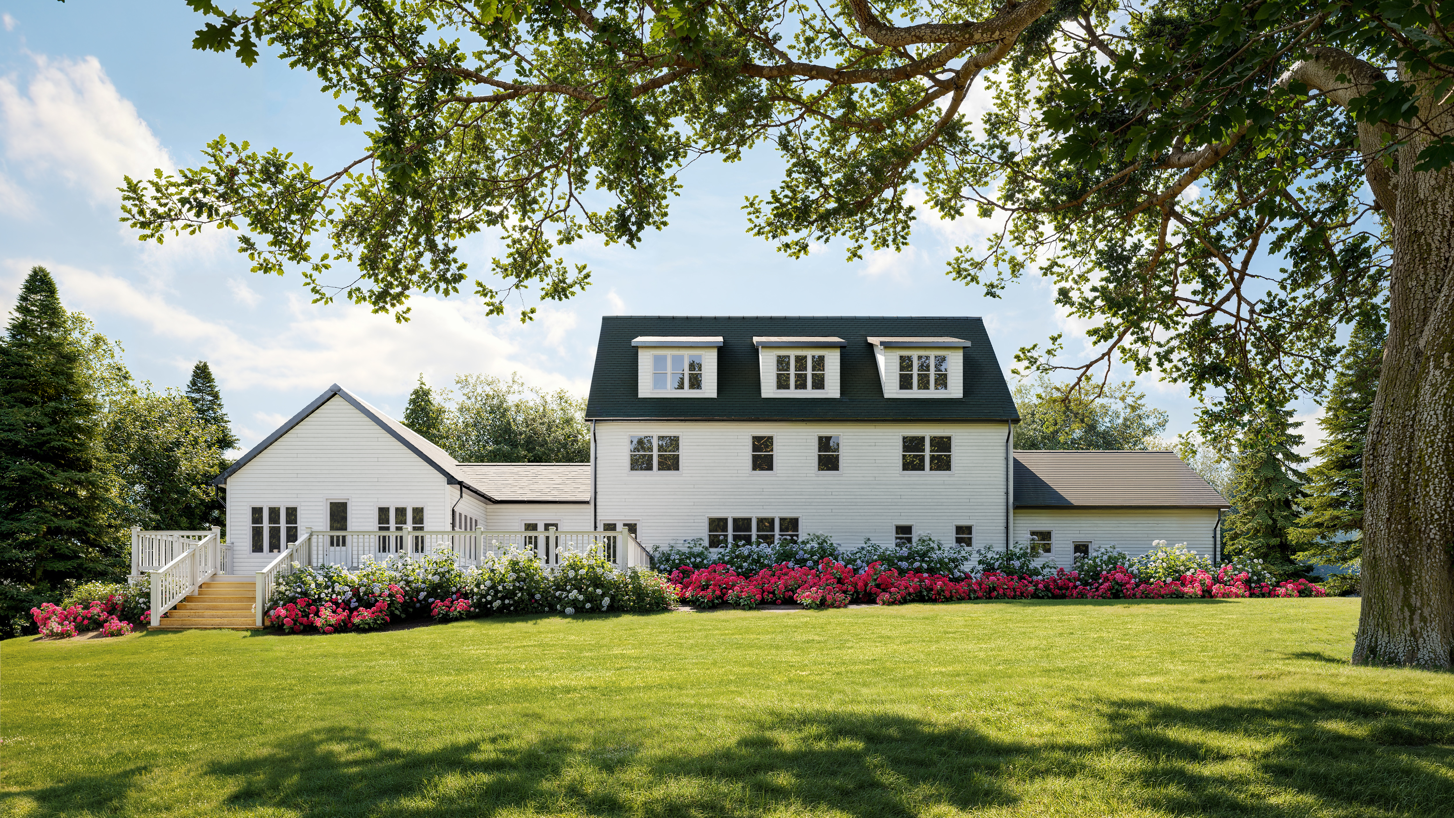 White farmhouse with dormers, surrounded by green lawn and colorful flower beds.